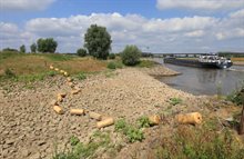 Droge rivierbedding met blootgelegde stenen bij lage waterstand, vrachtschip vaart voorbij.